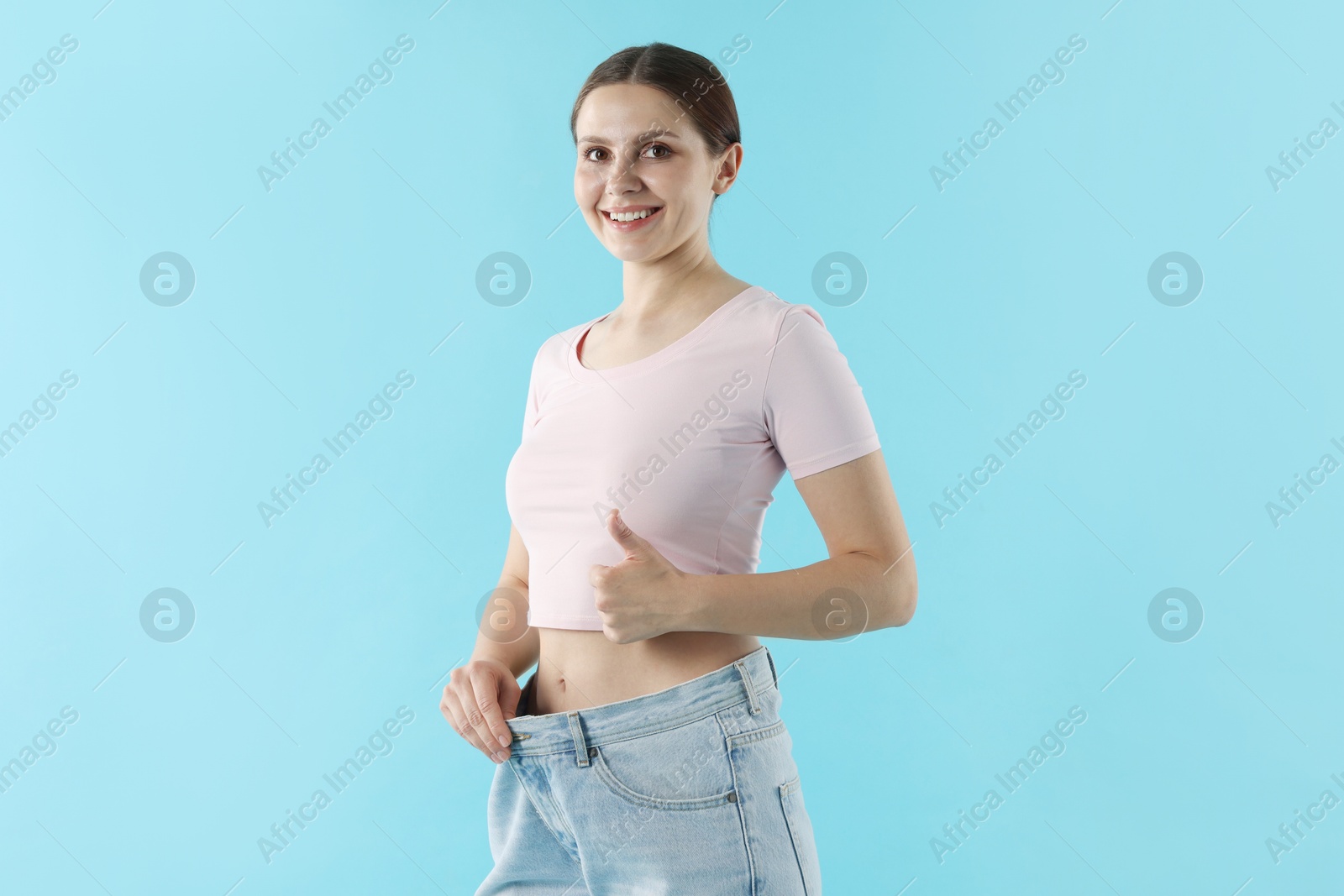 Photo of Beautiful woman wearing old jeans after weight loss and showing thumbs up on light blue background