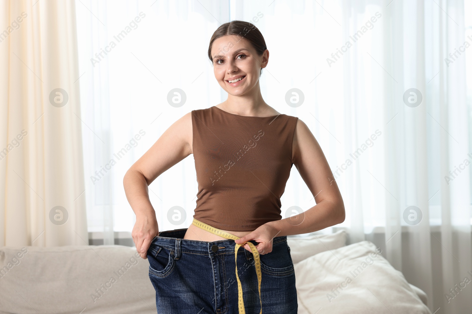 Beautiful woman with measuring tape wearing old jeans after weight loss indoors Photo of Beautiful woman with measuring tape wearing old jeans after weight loss indoors