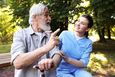 Photo of Smiling nurse supporting elderly man with walking cane on bench outdoors