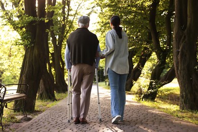 Photo of Caregiver supporting elderly man who using walking frame outdoors, back view
