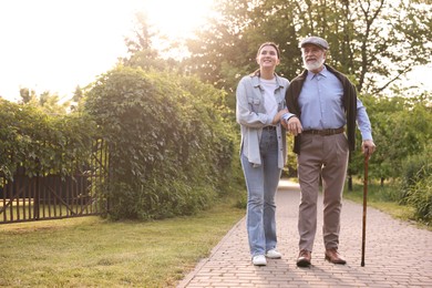 Photo of Smiling caregiver supporting elderly man with walking cane outdoors. Space for text