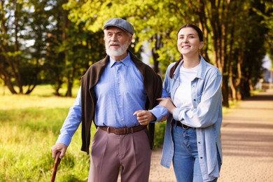 Photo of Smiling caregiver supporting elderly man with walking cane outdoors