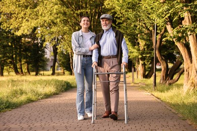 Photo of Smiling caregiver supporting elderly man who using walking frame outdoors
