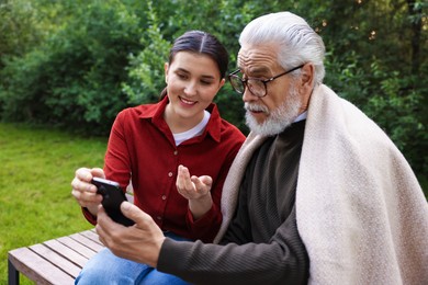 Photo of Smiling caregiver and elderly man in glasses using smartphone on bench outdoors