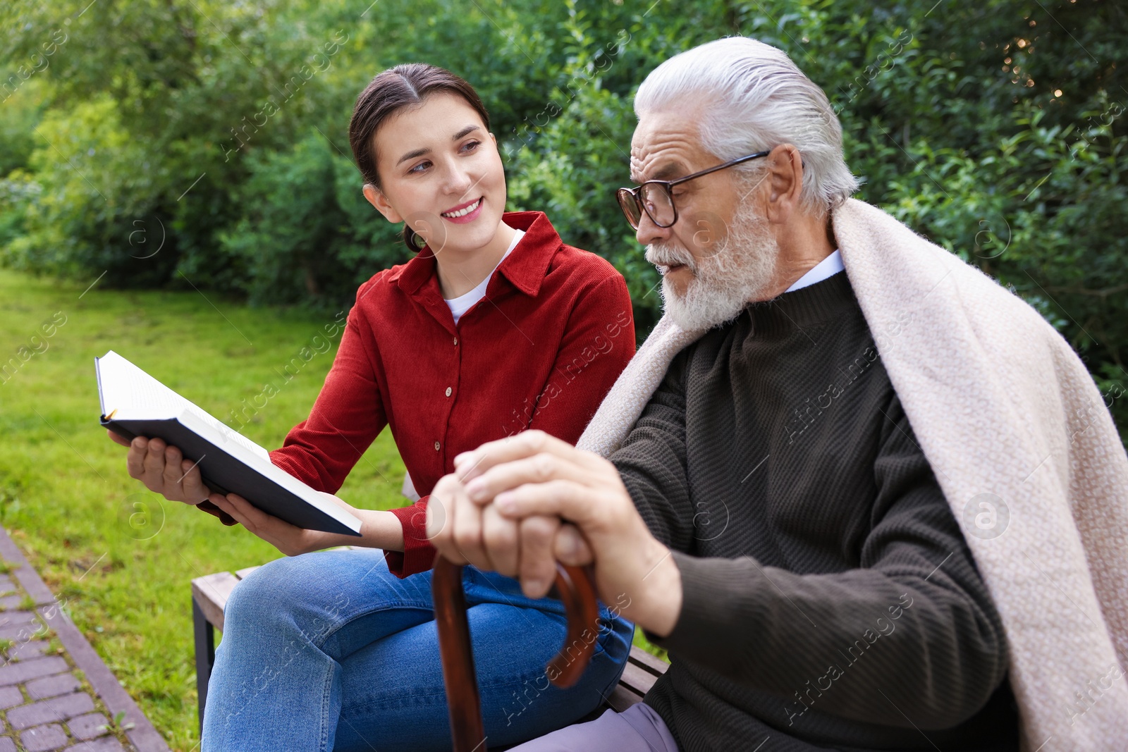 Smiling caregiver and elderly man in glasses with walking cane reading book on bench outdoors Photo of Smiling caregiver and elderly man in glasses with walking cane reading book on bench outdoors