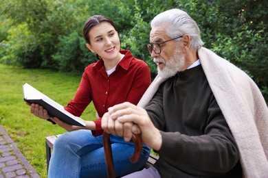 Photo of Smiling caregiver and elderly man in glasses with walking cane reading book on bench outdoors