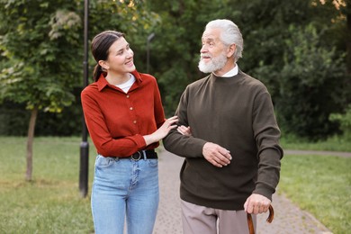Photo of Smiling caregiver supporting elderly man who using walking cane outdoors