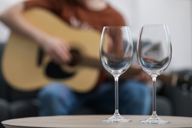 Photo of Empty wineglasses on table and man playing guitar indoors, selective focus