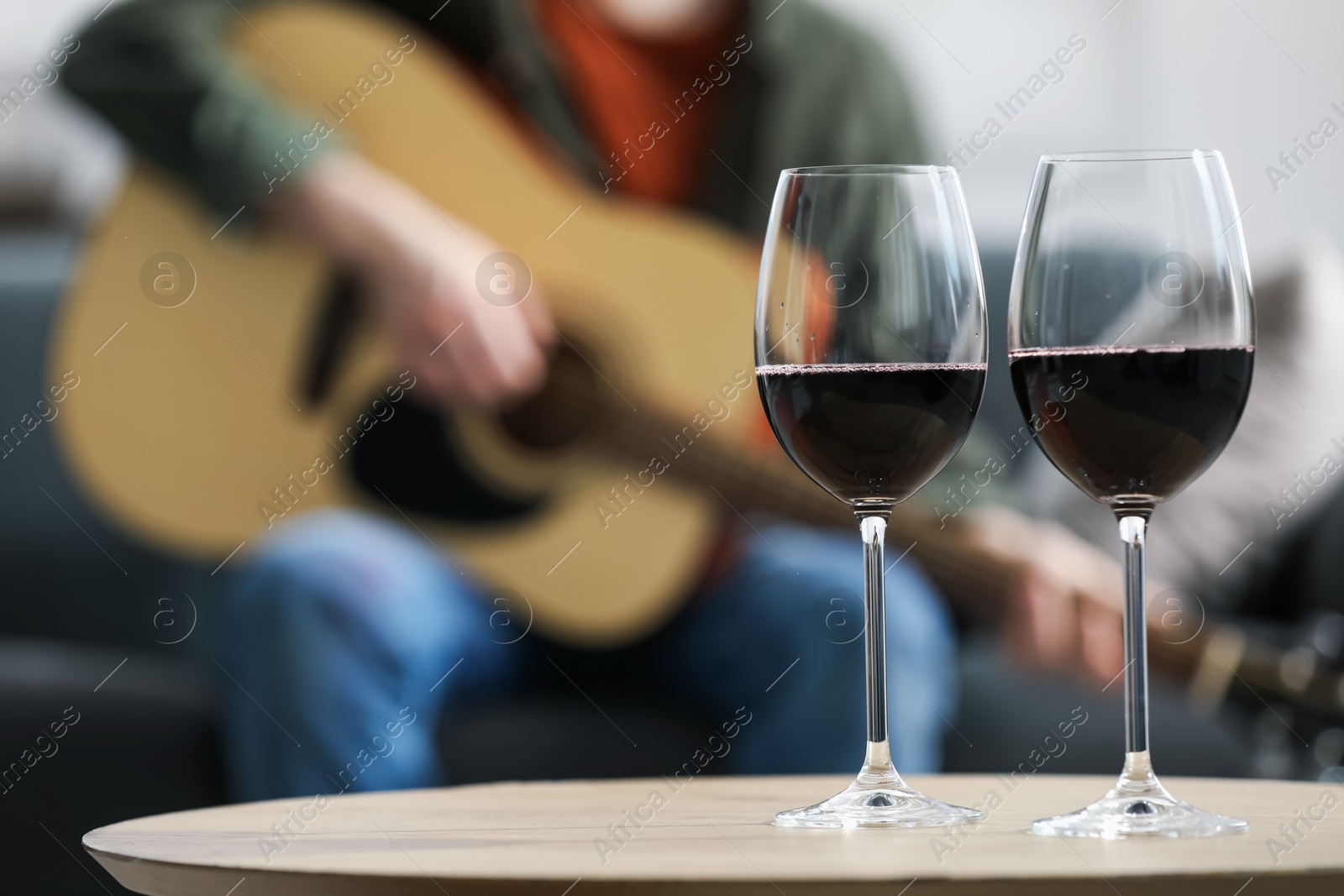 Glasses with red wine on table and man playing guitar indoors, selective focus Photo of Glasses with red wine on table and man playing guitar indoors, selective focus