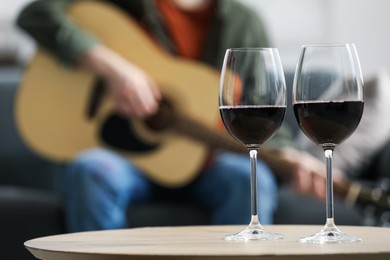 Photo of Glasses with red wine on table and man playing guitar indoors, selective focus