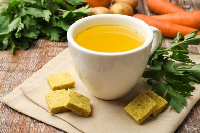 Photo of Aromatic bouillon cubes, broth, parsley and vegetables on wooden table, closeup