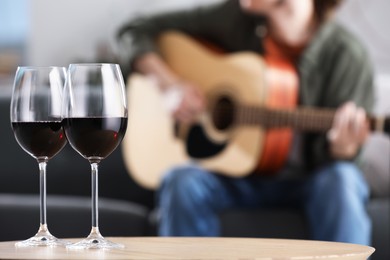 Photo of Glasses with red wine on table and man playing guitar indoors, selective focus