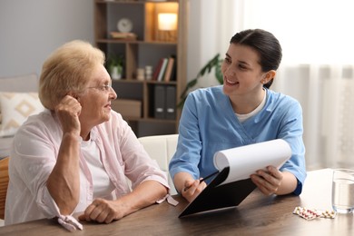 Photo of Nurse with clipboard examining senior woman at wooden table indoors. Home health care service
