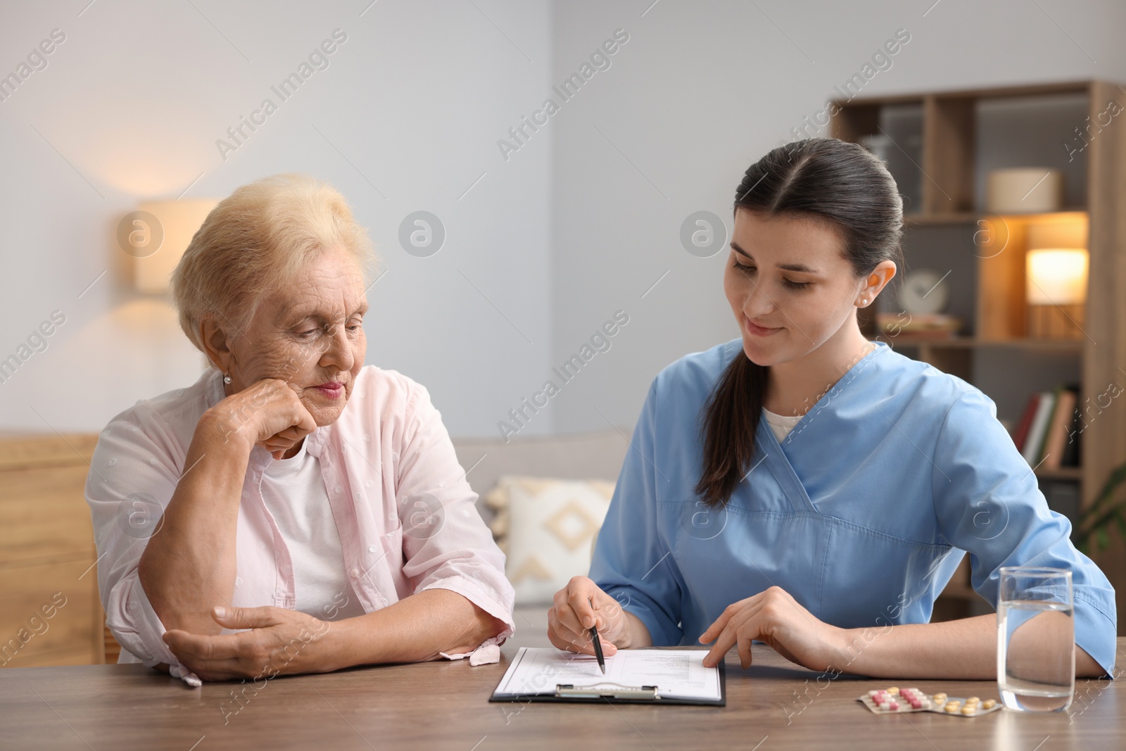 Nurse with clipboard examining senior woman at wooden table indoors. Home health care service Photo of Nurse with clipboard examining senior woman at wooden table indoors. Home health care service