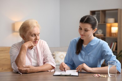 Photo of Nurse with clipboard examining senior woman at wooden table indoors. Home health care service