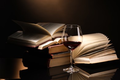 Photo of Red wine in glass and stack of books on mirror table against dark background, closeup