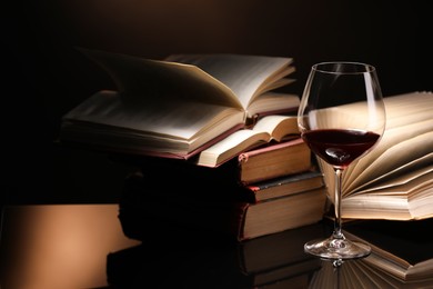 Photo of Red wine in glass and stack of books on mirror table against dark background, closeup