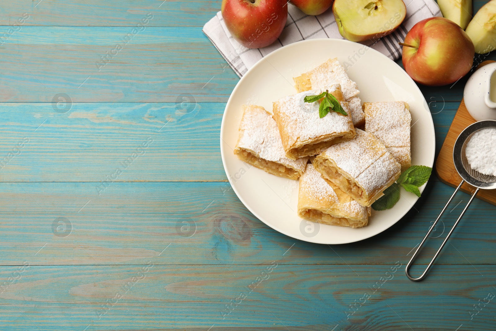 Pieces of tasty apple strudel with powdered sugar, mint and fruits on light blue wooden table, flat lay. Space for text Photo of Pieces of tasty apple strudel with powdered sugar, mint and fruits on light blue wooden table, flat lay. Space for text