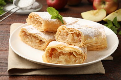 Photo of Pieces of tasty apple strudel with powdered sugar and mint on wooden table, closeup