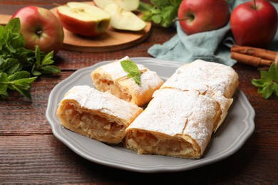 Photo of Pieces of tasty apple strudel with powdered sugar, mint and fruits on wooden table, closeup