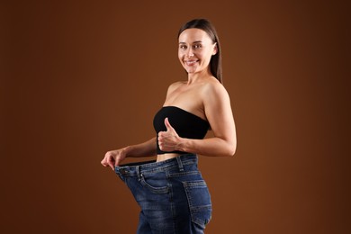 Photo of Fit woman wearing old jeans after weight loss and showing thumbs up on brown background