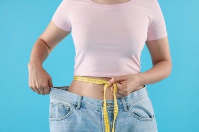 Photo of Woman with measuring tape wearing old jeans after weight loss on light blue background, closeup