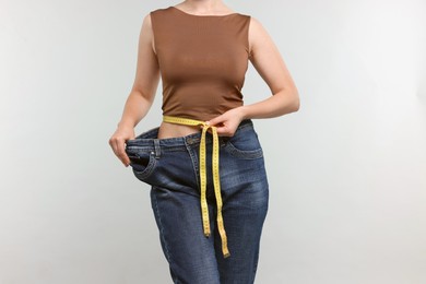 Photo of Woman with measuring tape wearing old jeans after weight loss on light grey background, closeup