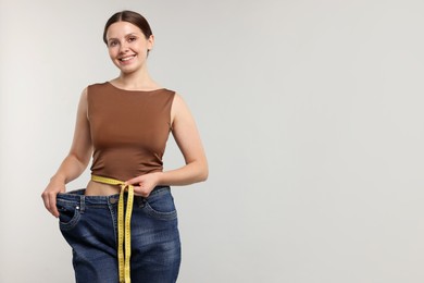 Photo of Beautiful woman with measuring tape wearing old jeans after weight loss on light grey background, space for text