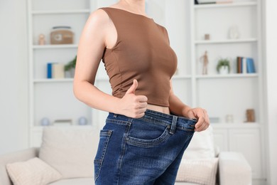 Photo of Woman wearing old jeans after weight loss and showing thumbs up indoors, closeup