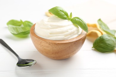Photo of Delicious yogurt in bowl, basil and spoon on white wooden table, closeup