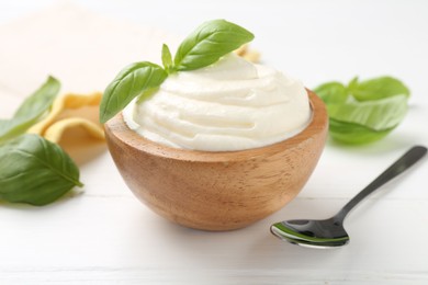 Photo of Delicious yogurt in bowl, basil and spoon on white wooden table, closeup