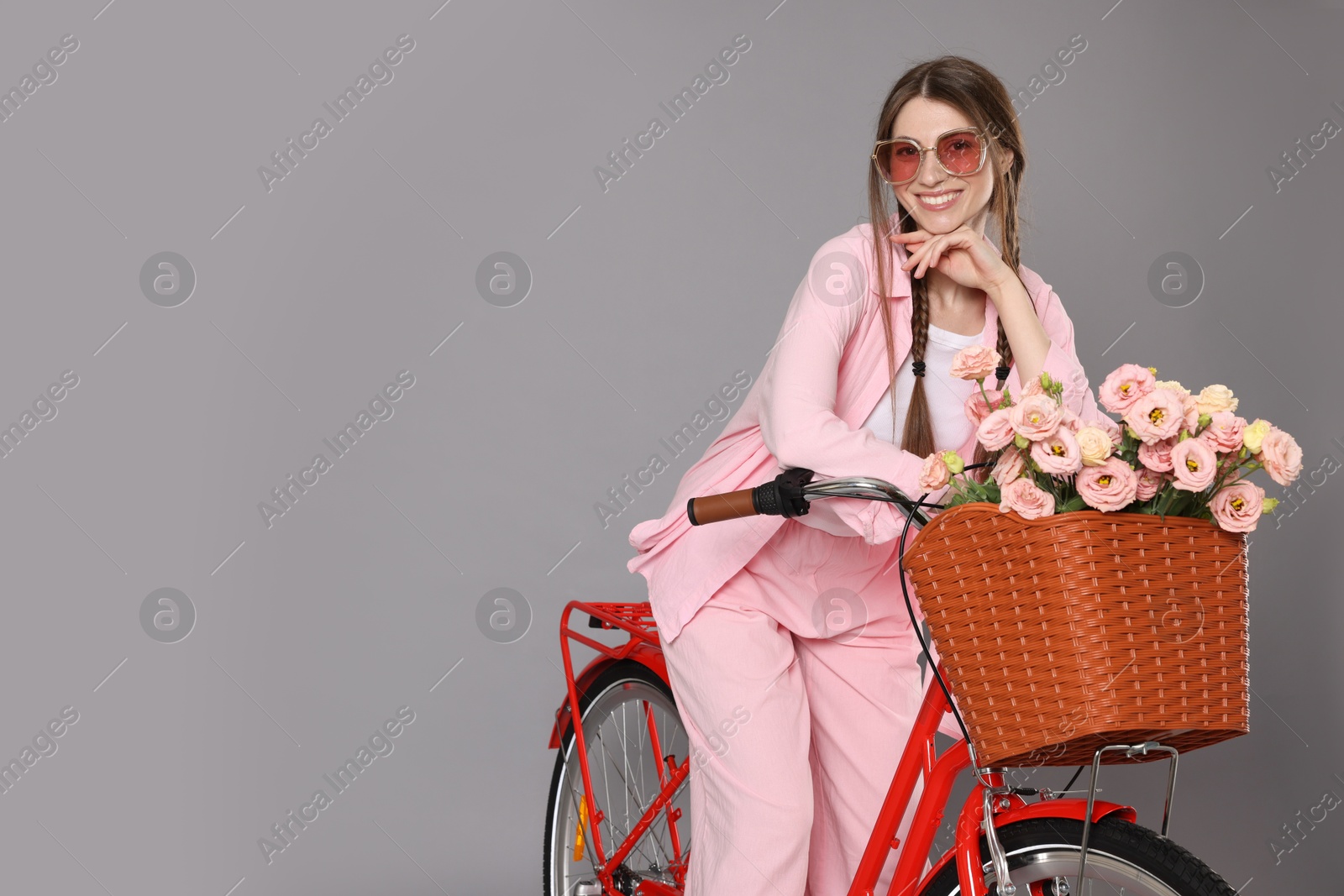 Smiling woman in sunglasses posing on bicycle with basket of flowers against grey background. Space for text Photo of Smiling woman in sunglasses posing on bicycle with basket of flowers against grey background. Space for text