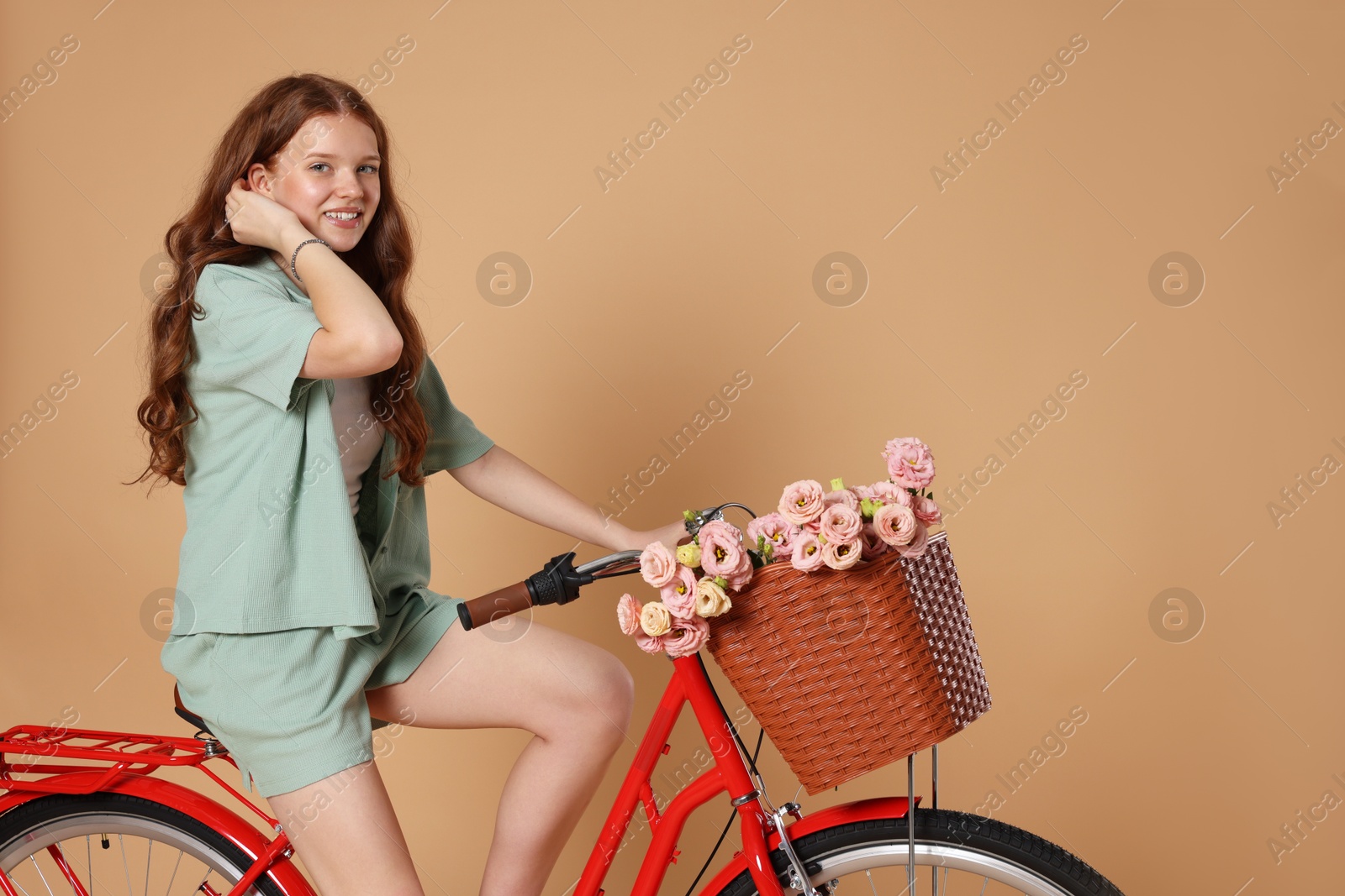 Smiling teenage girl riding bicycle with basket of flowers against beige background Photo of Smiling teenage girl riding bicycle with basket of flowers against beige background