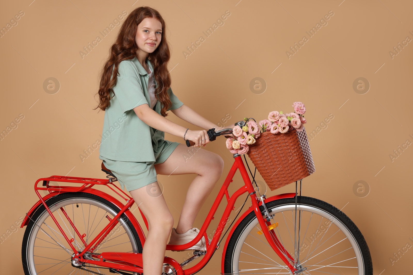 Beautiful teenage girl riding bicycle with basket of flowers against beige background Photo of Beautiful teenage girl riding bicycle with basket of flowers against beige background
