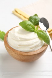 Photo of Delicious yogurt in bowl, basil and spoon on white wooden table, closeup