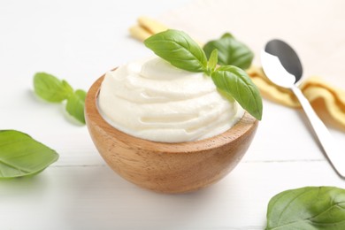 Photo of Delicious yogurt in bowl, basil and spoon on white wooden table, closeup