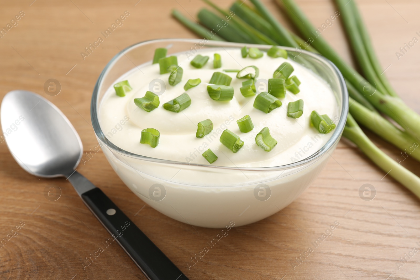Photo of Delicious yogurt in bowl, green onions and spoon on wooden table, closeup