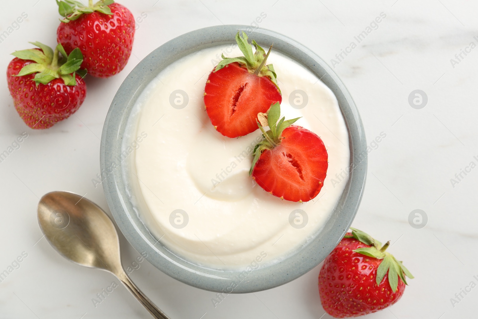 Photo of Delicious yogurt in bowl, strawberries and spoon on white marble table, flat lay