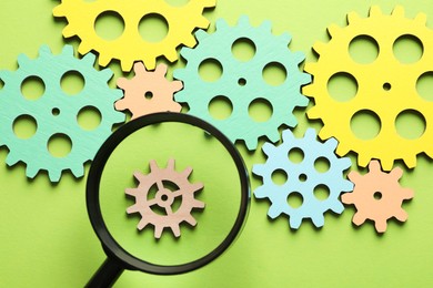 Photo of Unemployment. Gearwheels and magnifying glass on green background, flat lay
