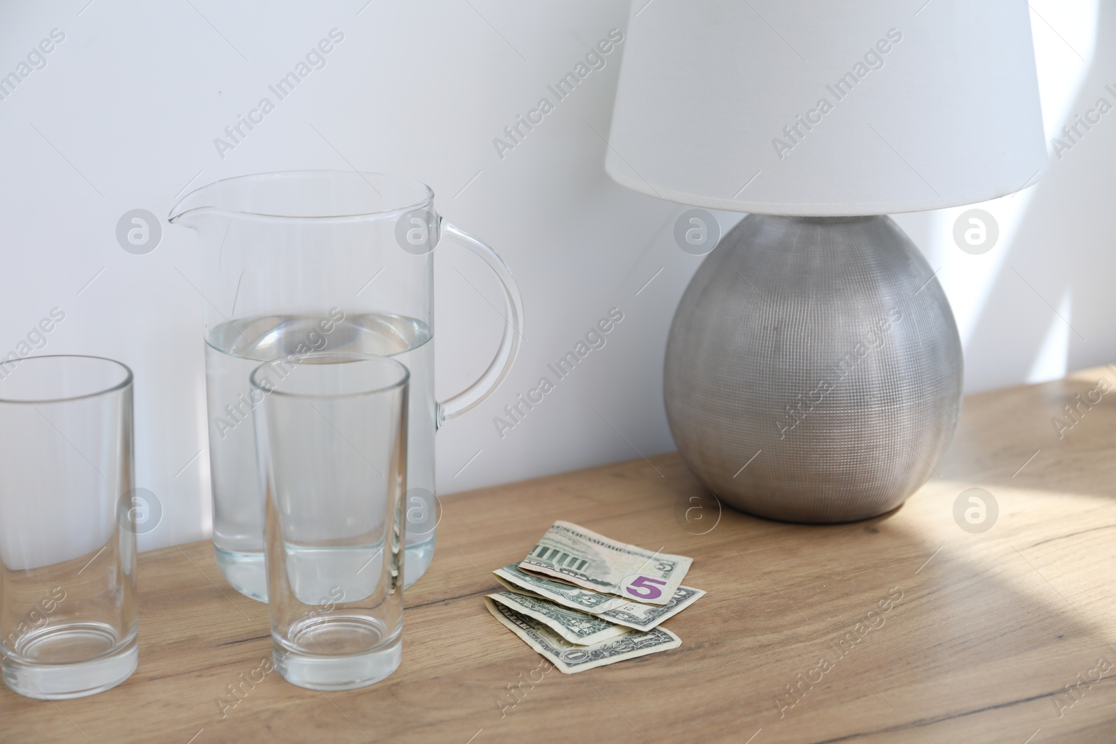 Photo of Tips, glasses and jug of water on wooden nightstand in hotel room, closeup