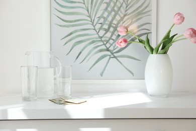 Photo of Tips, flowers, glasses and jug of water on white nightstand in hotel room