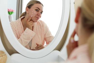 Photo of Woman applying cream onto face near mirror indoors. Acne treatment