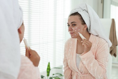 Photo of Woman applying cream onto face near mirror indoors. Acne treatment