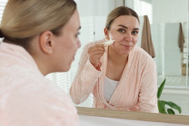Photo of Woman applying cream onto face near mirror indoors. Acne treatment