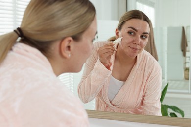 Photo of Woman applying cream onto face near mirror indoors. Acne treatment
