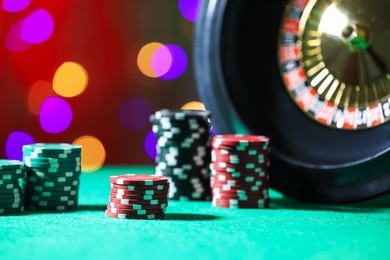 Photo of Casino chips and roulette wheel on green table against blurred lights, closeup