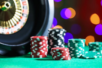 Photo of Casino chips and roulette wheel on green table against blurred lights, closeup