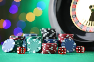 Photo of Casino chips, dice and roulette wheel on green table against blurred lights, closeup