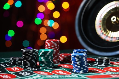 Photo of Casino chips and roulette wheel on table against blurred lights, closeup