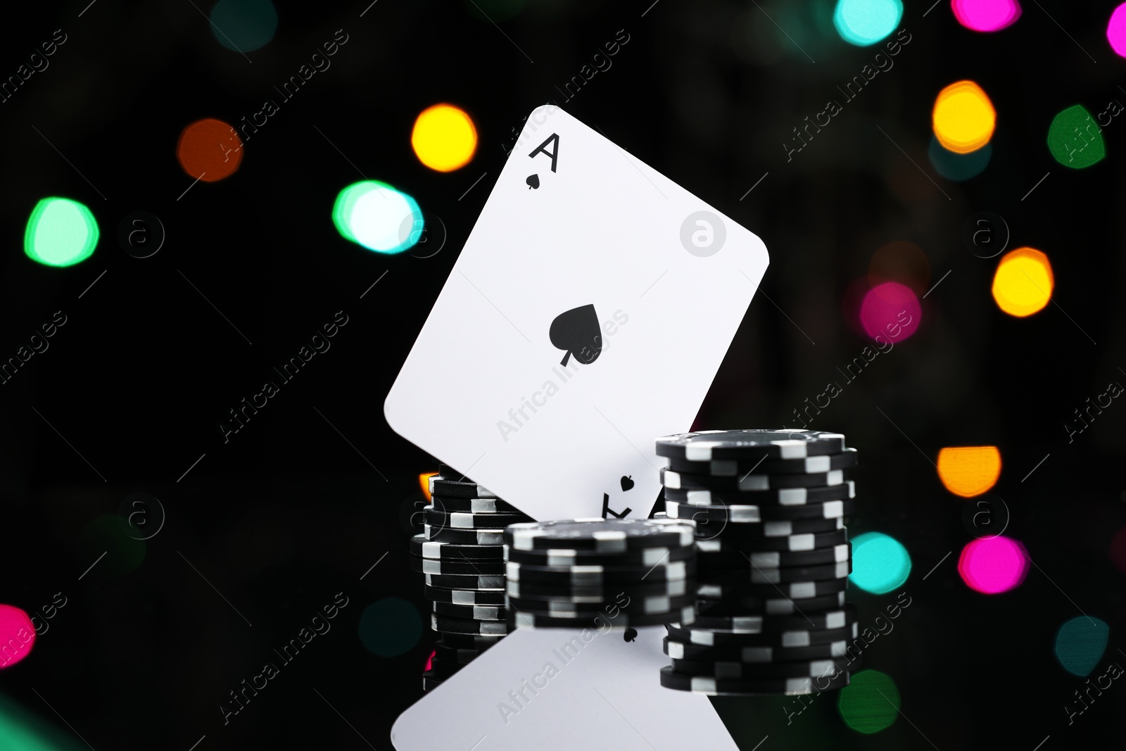 Photo of Casino chips and playing card on mirror surface against blurred lights, closeup